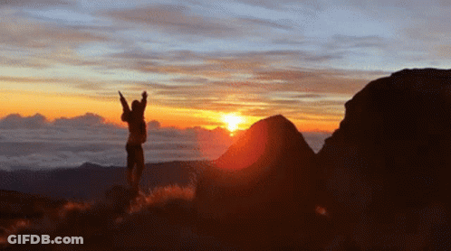 A Person Is Standing On Top Of A Hill With Their Arms In The Air At Sunset GIF