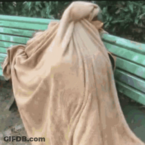 A Person Is Wrapped In A Blanket While Sitting On A Green Bench . GIF
