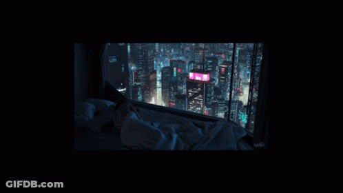 A Person Laying On A Bed Looking Out A Window At The City At Night GIF
