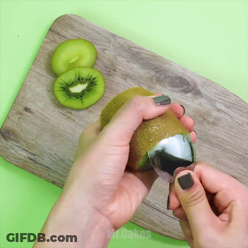 A Person Peeling A Kiwi With A Spoon On A Wooden Cutting Board GIF