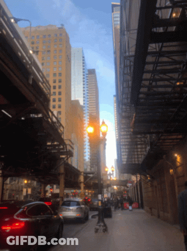 A Person Pushing A Stroller Down A City Street With Tall Buildings In The Background GIF