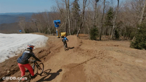 A Person Riding A Bike Down A Dirt Road With A Snow Cannon In The Background GIF