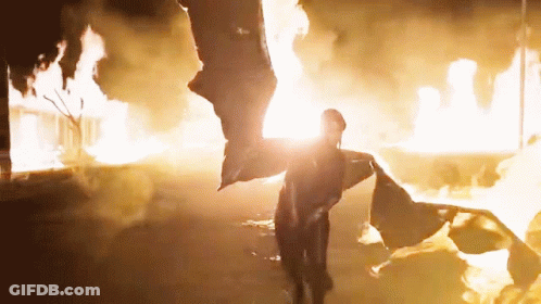 A Person Standing In Front Of A Fire With A Flag Flying In The Air GIF