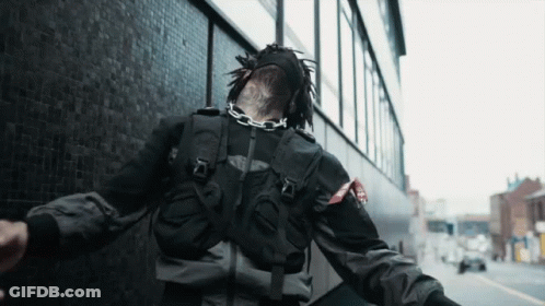 A Person Wearing A Black Jacket And A Mask With A Chain Around Their Neck Is Walking Down The Street GIF