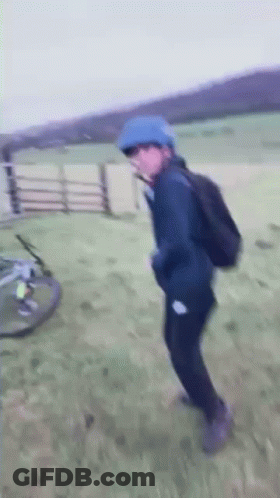 A Person Wearing A Blue Helmet And A Backpack Is Standing In A Field With A Bicycle In The Background . GIF
