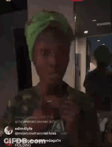 A Person Wearing A Green Hat Is Standing In A Room With A Holding Sign In The Corner GIF