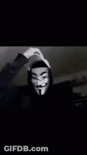 A Person Wearing A Hooded Jacket And A Anonymous Mask Is Waving At The Camera . GIF