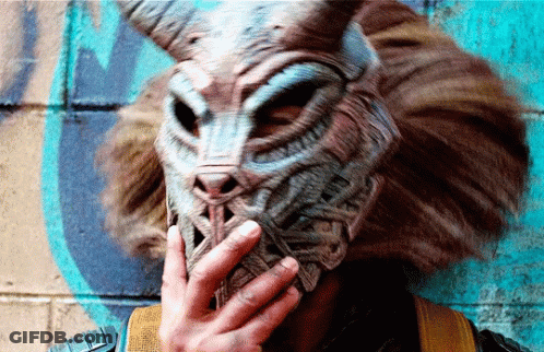 A Person Wearing A Mask With Horns Is Covering Their Mouth With Their Hand GIF