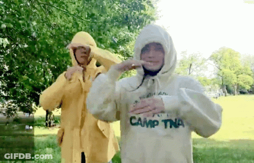 A Person Wearing A White Camp Tna Sweatshirt Stands Next To Another Person Wearing A Yellow Raincoat GIF