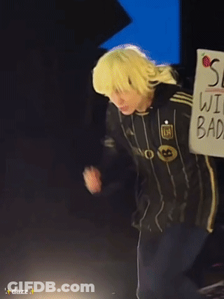 A Person Wearing A Yellow Wig And A Black Jacket With The Letter M On It GIF