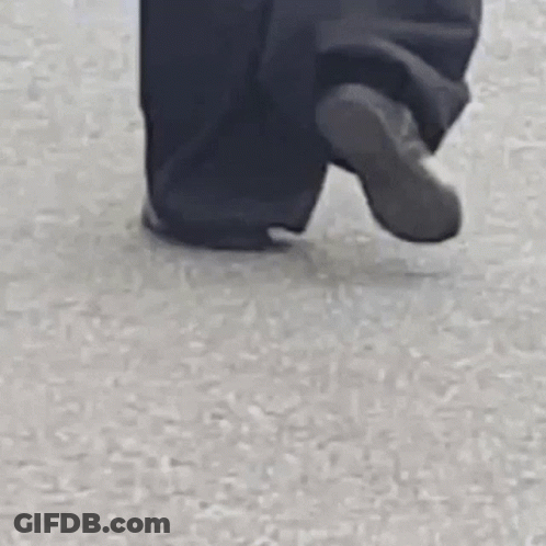 A Person Wearing Black Pants And A White Jacket Is Walking On A Street . GIF