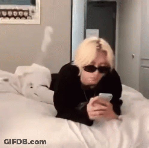 A Person Wearing Sunglasses Is Laying On A Bed Looking At Their Phone . GIF