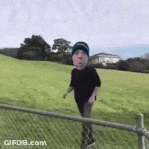 A Person With A Face On Their Head Is Walking Through A Grassy Field Behind A Chain Link Fence . GIF