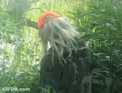 A Person With Long Blonde Hair And An Orange Hat Stands In The Woods GIF