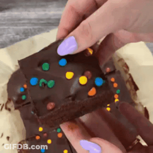 A Person With Purple Nails Is Holding A Piece Of Chocolate Cake With Sprinkles On Top . GIF