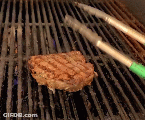 A Piece Of Meat Is Being Cooked On A Grill With Tongs GIF
