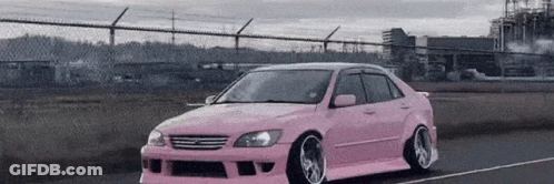 A Pink Car Is Driving Down A Road Next To A Chain Link Fence . GIF