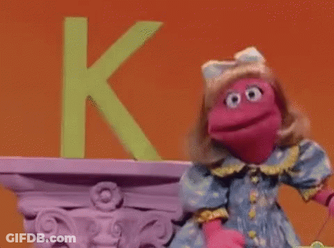 A Pink Puppet Is Standing In Front Of A Green Letter K On A Shelf . GIF