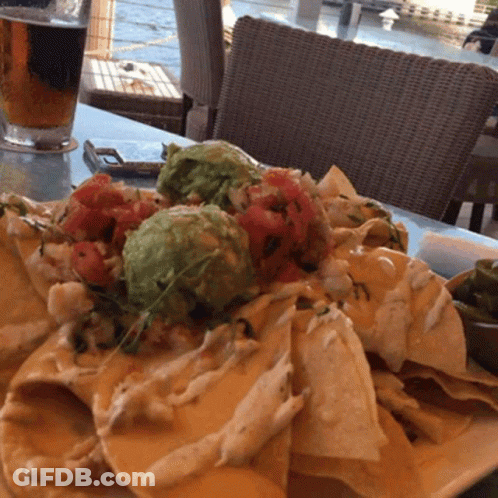 A Plate Of Nachos Sits On A Table With A Glass Of Beer In The Background GIF