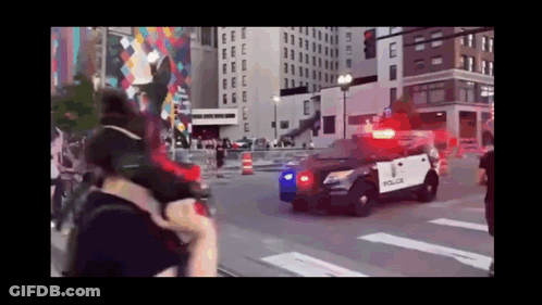 A Police Car Is Driving Down A City Street With A Crowd Behind It . GIF