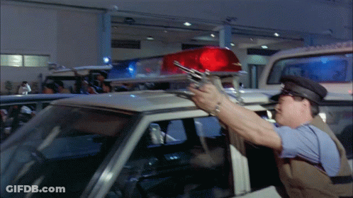 A Police Officer Holds A Gun In Front Of A Police Car GIF