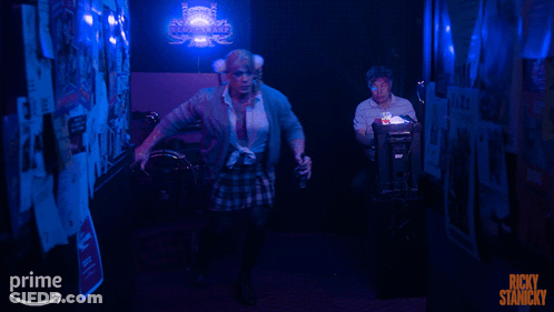 A Poster For Ricky Stanicky Shows A Man In A Plaid Skirt In A Dark Hallway GIF
