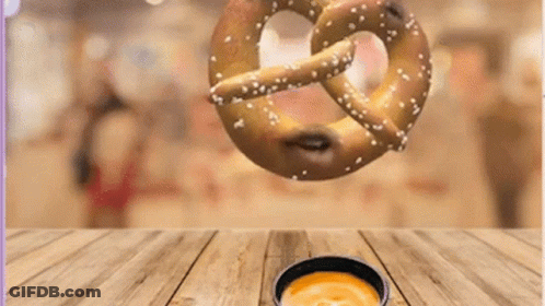 A Pretzel Is Falling Into A Bowl Of Cheese Sauce On A Wooden Table GIF