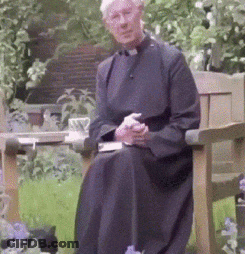 A Priest Is Sitting On A Bench In A Garden With A Cat GIF