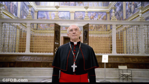 A Priest With A Cross Around His Neck Stands In Front Of A Building GIF
