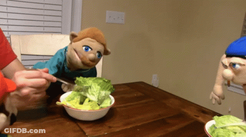 A Puppet Is Eating Lettuce From A Bowl On A Table GIF