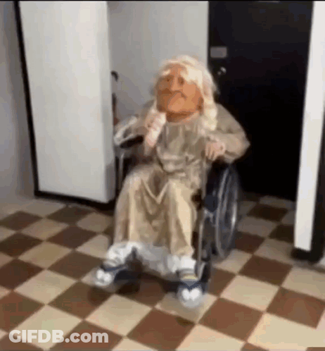 A Puppet Is Sitting In A Wheelchair On A Checkered Floor In A Hallway . GIF