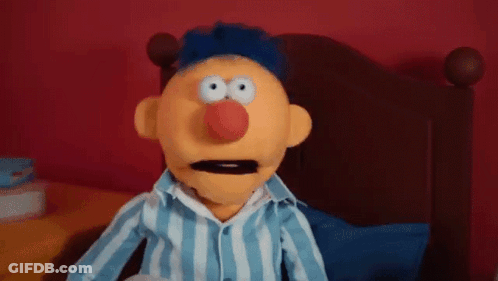 A Puppet Is Sitting On A Bed Wearing A Blue And White Striped Shirt And A Red Nose . GIF