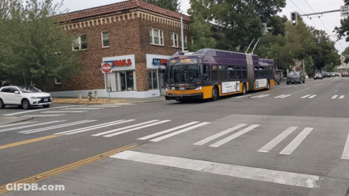 A Purple Bus Is Driving Down The Street In Front Of A Building That Says Nails GIF