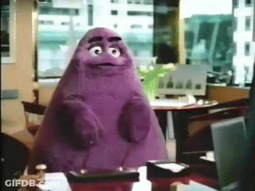 A Purple Cartoon Character Is Sitting At A Table In An Office GIF