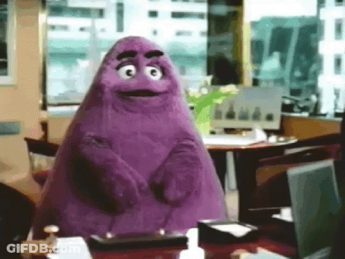 A Purple Cartoon Character Is Standing In Front Of A Desk With A Laptop On It . GIF