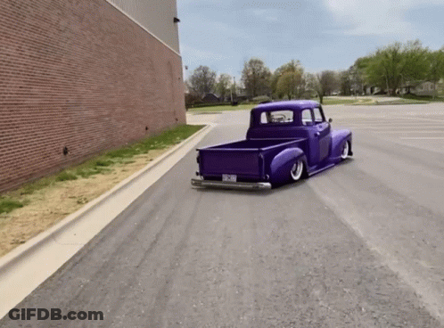 A Purple Truck Is Driving Down A Street With A Brick Building In The Background And A Few Trees GIF