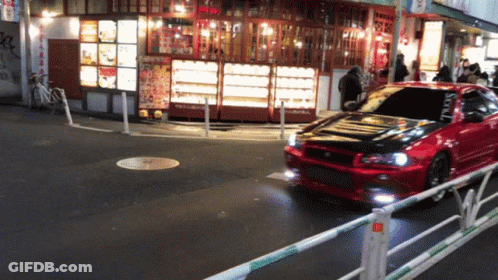 A Red Car Is Driving Down A Street In Front Of A Restaurant With A Sign That Says ' Noodles ' On It GIF