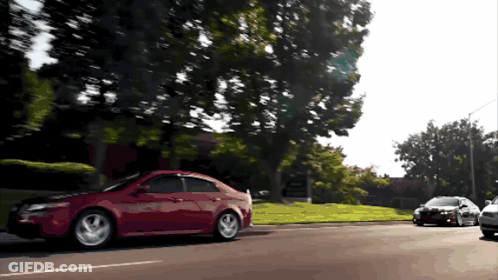 A Red Car Is Driving Down A Street Next To A Black Car GIF