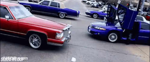 A Red Car Is Driving Down A Street Next To A Blue Car With Its Hood Open GIF