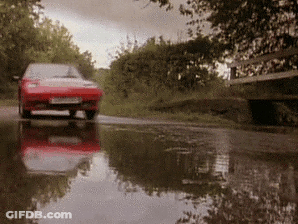 A Red Car Is Driving Through A Puddle On A Wet Road GIF