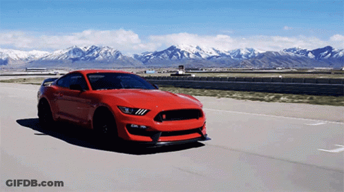 A Red Ford Mustang Is Driving Down A Road With Mountains In The Background GIF