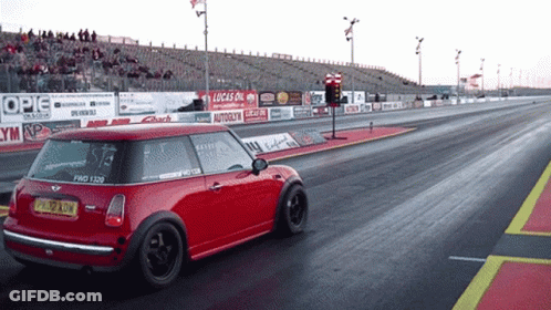 A Red Mini Cooper Is Driving Down A Race Track With A Lucas Oil Sign Behind It GIF