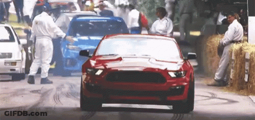 A Red Mustang Is Driving Down A Street With A Crowd Of People Behind It . GIF