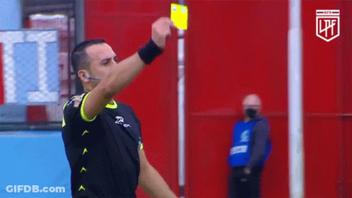 A Referee Holds Up A Yellow Card In Front Of A Red Wall With The Letters Lpf On It GIF