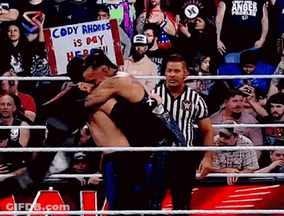 A Referee Is Standing In A Wrestling Ring Holding A Sign That Says `` Cody Rhodes Is My Hero '' GIF