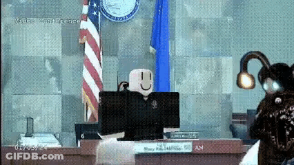A Roblox Character Is Sitting At A Desk In Front Of A Computer In A Courtroom With An American Flag In The Background . GIF