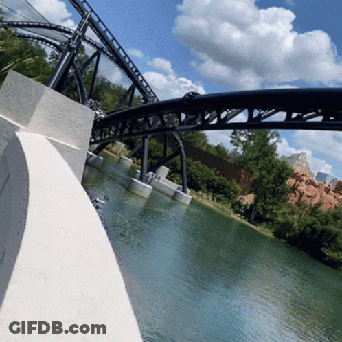A Roller Coaster Going Over A Body Of Water With Trees In The Background GIF