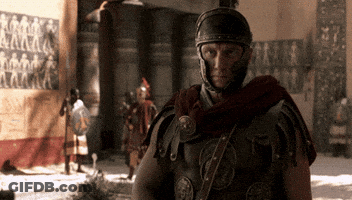 A Roman Soldier With A Helmet On Stands In Front Of A Wall With Images Of People On It GIF