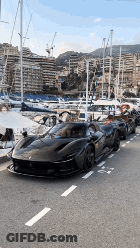 A Row Of Black Sports Cars Are Parked On The Side Of The Road Near A Marina GIF
