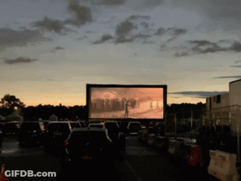 A Row Of Cars Are Parked In Front Of A Large Screen That Shows A Movie GIF